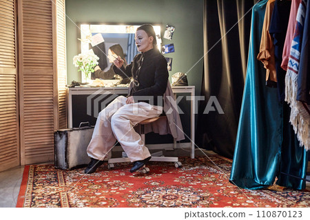 Full length portrait of female mime performer sitting by mirror backstage preparing for show in theater, copy space Full length portrait of female mime performer sitting by mirror backstage preparing for show in theater, copy space 110870123