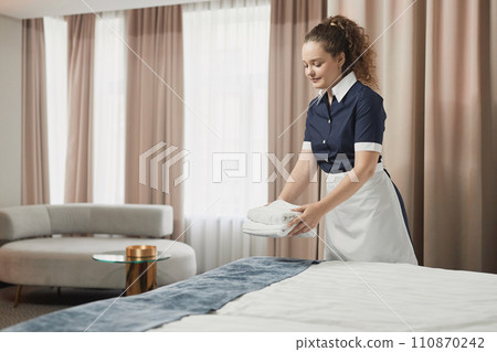 Portrait of smiling young housekeeper setting up fresh towels on bed in hotel room and preparing for guests, copy space Portrait of smiling young housekeeper setting up fresh towels on bed in hotel room and preparing for guests, copy space 110870242