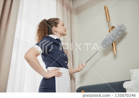 Side view portrait of smiling young maid dusting light fixtures on wall while cleaning hotel room and preparing for guests, copy space 110870246