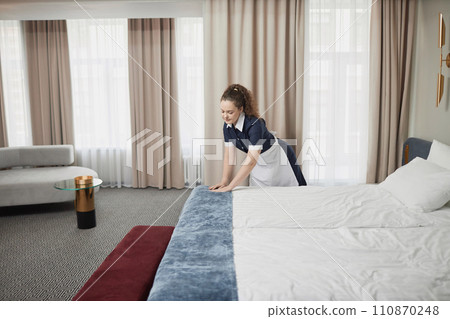 Wide angle view at young housekeeper cleaning hotel room and making comfortable bed with white sheets preparing for guests, copy space 110870248