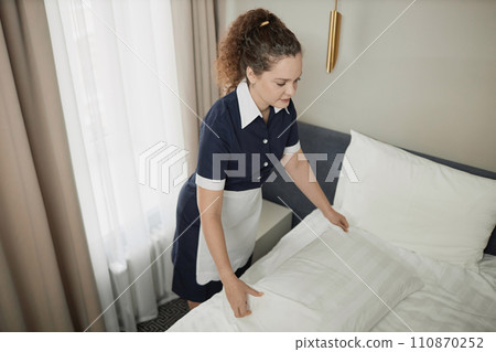 High angle view of young maid wearing uniform making bed with white sheets while cleaning hotel room, copy space High angle view of young maid wearing uniform making bed with white sheets while cleaning hotel room, copy space 110870252