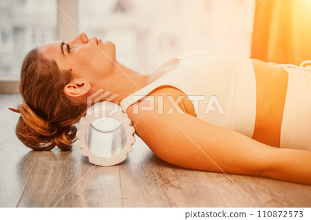 Adult athletic woman, in white bodysuit, performing fascia exercises on the floor - caucasian woman using a massage foam roller - a tool to relieve tension in the back and relieve muscle pain - the 110872573
