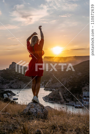 Happy woman standing with her back on the sunset in nature in summer with open hands posing with mountains on sunset, silhouette. Woman in the mountains red dress, eco friendly, summer landscape 110872639
