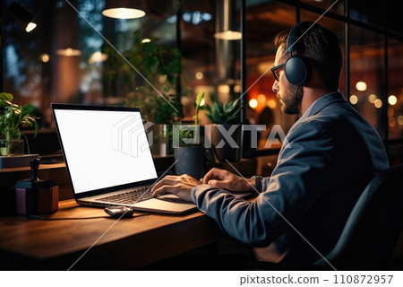 Man works in the evening at a laptop with overhead headphones 110872957