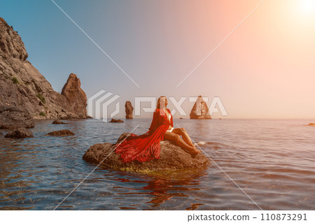 woman sea red dress. Beautiful sensual woman in a flying red dress and long hair, sitting on a rock above the beautiful sea in a large bay. woman sea red dress. Beautiful sensual woman in a flying red dress and long hair, sitting on a rock above the beautiful sea in a large bay. 110873291