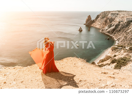 Woman red dress sea. Female dancer in a long red dress posing on a beach with rocks on sunny day. Girl on the nature on blue sky background. Woman red dress sea. Female dancer in a long red dress posing on a beach with rocks on sunny day. Girl on the nature on blue sky background. 110873297