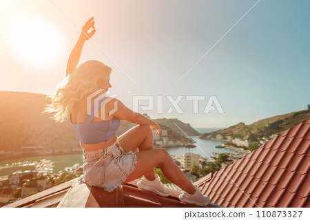 Woman sits on rooftop with outstretched arms, enjoys town view and sea mountains. Peaceful rooftop relaxation. Below her, there is a town with several boats visible in the water Woman sits on rooftop with outstretched arms, enjoys town view and sea mountains. Peaceful rooftop relaxation. Below her, there is a town with several boats visible in the water 110873327