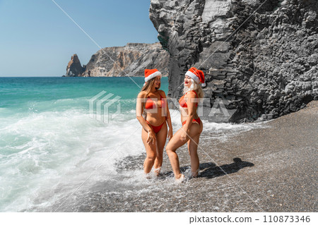 Women in Santa Claus hats run into the sea dressed in red swimsuits. Celebrating the New Year in a hot country Women in Santa Claus hats run into the sea dressed in red swimsuits. Celebrating the New Year in a hot country 110873346