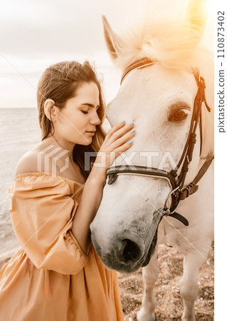 A woman in a dress stands next to a white horse on a beach, with the blue sky and sea in the background. A woman in a dress stands next to a white horse on a beach, with the blue sky and sea in the background. 110873402