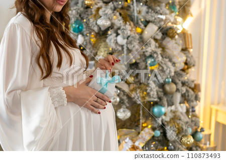Pregnant woman holds a gift on her stomach against Christmas tree with lights. family holiday concept Christmas holidays. 110873493