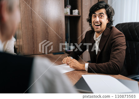 Portrait of cheerful male boss in suit telling funny joke, having fun talking with employees during group briefing. Friendly business team laughing at table. Positive conversation engaged in teamwork 110873503