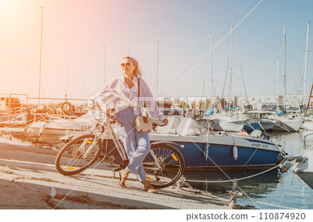Woman enjoys bike ride along waterfront, Marina surroundings. She is wearing a white shirt and blue jeans, and she has a handbag with her. Capturing outdoor bike ride by waterfront. 110874920