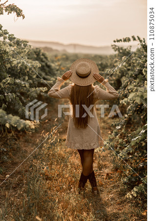 Woman with straw hat stands in front of vineyard. She is wearing a light dress and posing for a photo. Travel concept to different countries Woman with straw hat stands in front of vineyard. She is wearing a light dress and posing for a photo. Travel concept to different countries 110875034