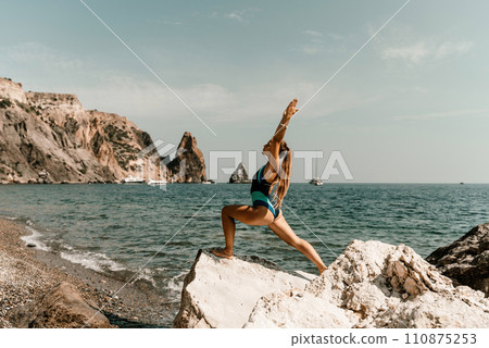 Yoga on the beach. A happy woman meditating in a yoga pose on the beach, surrounded by the ocean and rock mountains, promoting a healthy lifestyle outdoors in nature, and inspiring fitness concept. 110875253