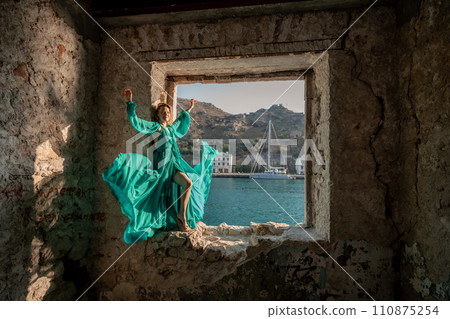 Rear view of a happy blonde woman in a long mint dress posing against the backdrop of the sea in an old building with columns. Girl in nature against the blue sky. Rear view of a happy blonde woman in a long mint dress posing against the backdrop of the sea in an old building with columns. Girl in nature against the blue sky. 110875254