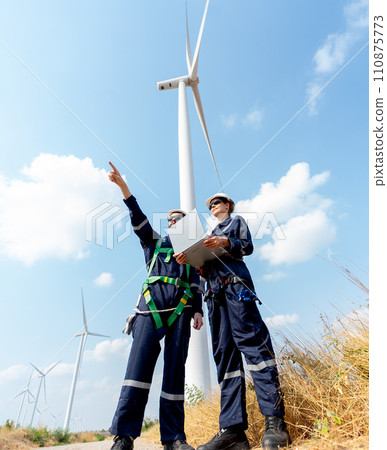 Vertical image of professional worker woman hold laptop and stand with her co-worker man point forward and they stand in front of wind turbine or windmill in the field for power plant business. Vertical image of professional worker woman hold laptop and stand with her co-worker man point forward and they stand in front of wind turbine or windmill in the field for power plant business. 110875773