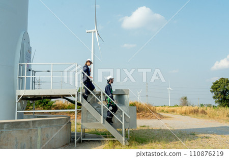 Two of technician workers walk down from stair of base of wind turbine or windmill after finish maintenance work in area of power plant. 110876219