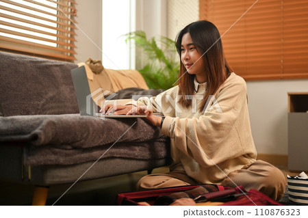 Young woman sitting in living room packing suitcase and using laptop for flight booking Young woman sitting in living room packing suitcase and using laptop for flight booking 110876323