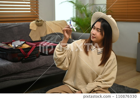 Young woman holding airplane model sitting near unpacked suitcases. Traveling, vacation and transportation concept Young woman holding airplane model sitting near unpacked suitcases. Traveling, vacation and transportation concept 110876330