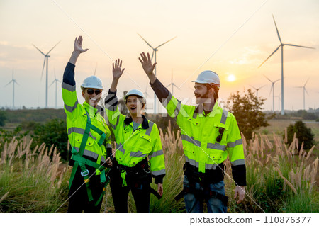 Group of technician worker man and woman stand with rest hands up express happiness during work and stand in front of windmill or wind turbine farm in area of power plant. Group of technician worker man and woman stand with rest hands up express happiness during work and stand in front of windmill or wind turbine farm in area of power plant. 110876377