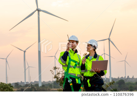 Professional technician man use walkie talkie to contact his co-worker and woman hold the laptop stand beside and work in front of windmill or wind turbine farm. 110876396
