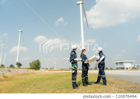 Group of professional technician workers stand with holding project paper plan in front of wind turbine or windmill and look back in area for power plant business. 110876494