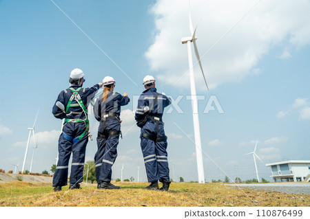 Back of group of professional technician man and woman workers with safety suit stand in front of wind turbine or windmill and point to the pole in area for power plant business. 110876499
