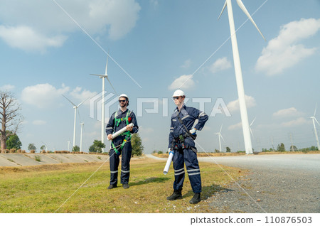 Professional Caucasian workers or technician with safety suit hold construction paper plan and stand in front of wind turbine or windmill in the row as power plant business. 110876503