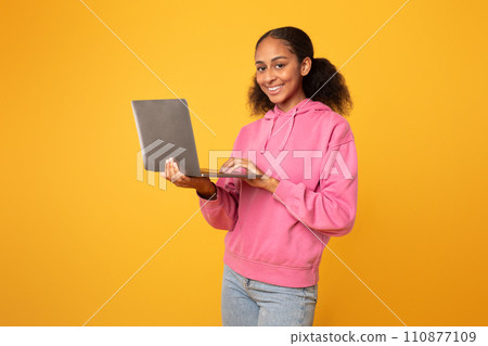 Cheerful black youngster girl with laptop browsing for homework, studio 110877109