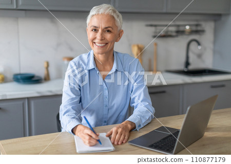 Cheerful mature lady sitting with laptop taking notes in kitchen 110877179