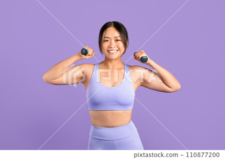 Happy Asian woman in sportswear with dumbbells on purple backdrop 110877200