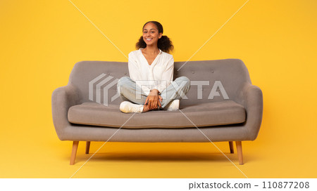Cheerful african american teen girl sitting on sofa, yellow backdrop Cheerful african american teen girl sitting on sofa, yellow backdrop 110877208