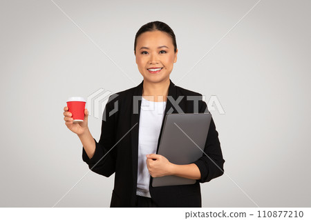 Professional Asian businesswoman with a friendly smile holding a red coffee cup and a grey laptop Professional Asian businesswoman with a friendly smile holding a red coffee cup and a grey laptop 110877210
