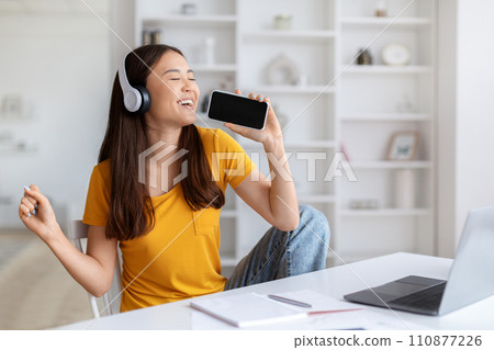 Asian woman with headphones joyfully singing while sitting at desk with laptop 110877226