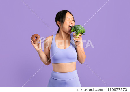 Asian woman in sportswear bites broccoli and holding donut 110877270