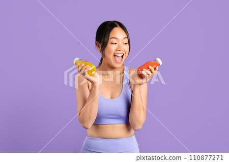 Excited Asian woman in sportswear holding smoothies on purple backdrop 110877271