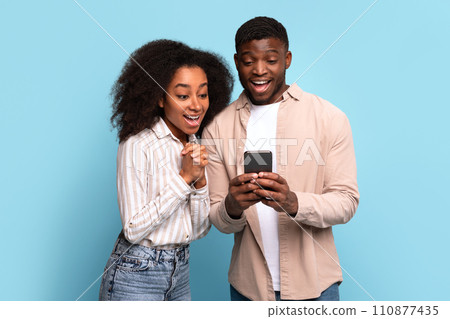 Excited black couple viewing smartphone together on blue background Excited black couple viewing smartphone together on blue background 110877435