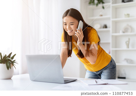 Asian woman having lively phone conversation and using laptop at home office 110877453