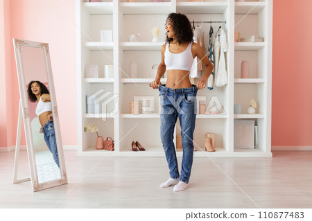 Joyful woman fitting into large jeans, standing by mirror 110877483