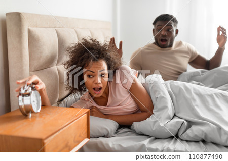 Shocked african spouses overslept looking at alarm clock in bedroom Shocked african spouses overslept looking at alarm clock in bedroom 110877490