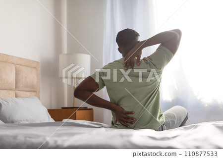 african man sitting on bed touching sore back and neck 110877533