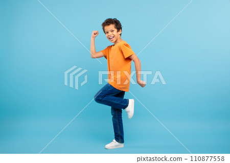 Joyful schoolboy in orange t-shirt clenching fist, blue background Joyful schoolboy in orange t-shirt clenching fist, blue background 110877558