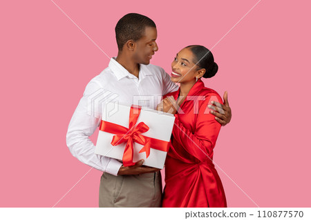 Smiling black man giving gift to grateful woman, both in elegant attire, pink backdrop 110877570