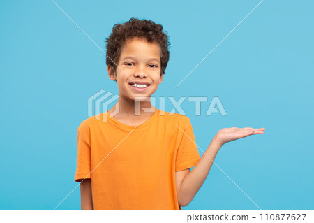 Happy boy presenting something with hand in orange shirt, blue background 110877627