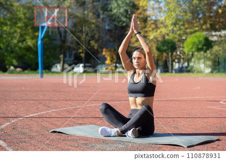 A concentrated young woman sitting on a yoga mat, performing a yoga pose confidently at an outdoor basketball court. A concentrated young woman sitting on a yoga mat, performing a yoga pose confidently at an outdoor basketball court. 110878931