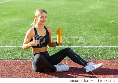 Smiling fit woman in sportswear sitting on a track field, giving a thumbs up with a water bottle in hand. Smiling fit woman in sportswear sitting on a track field, giving a thumbs up with a water bottle in hand. 110879119