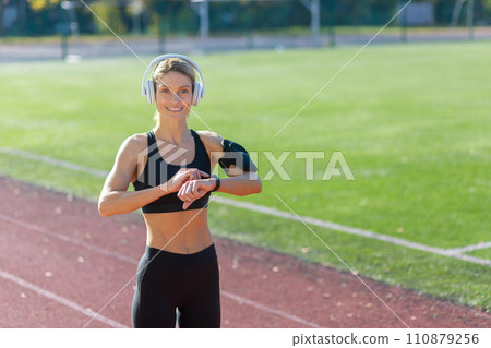 Active young woman wearing sportswear checks her smartwatch while enjoying music on her headphones at a track field. 110879256