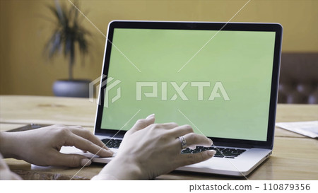 Young woman in pink shirt sitting at the large wooden table and typing on her laptop with chroma key green screen. Stock footage. Chroma key screen for placement of your own content. 110879356