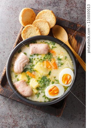 Polish soup zurek with white sausage and eggs close-up in a bowl. Vertical top view Polish soup zurek with white sausage and eggs close-up in a bowl. Vertical top view 110880013