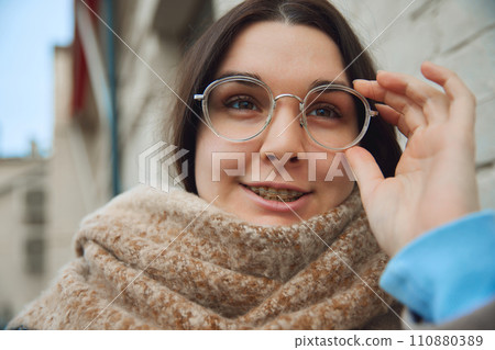 Close up portrait of young brunette woman dressed in warm outfit adjusts her stylish and fashionable glasses against blurred urban background. 110880389